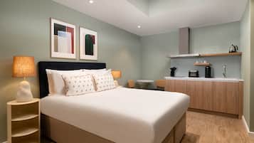 A modern hotel room with a large bed, bedside tables, a small round table, and a kitchenette area.