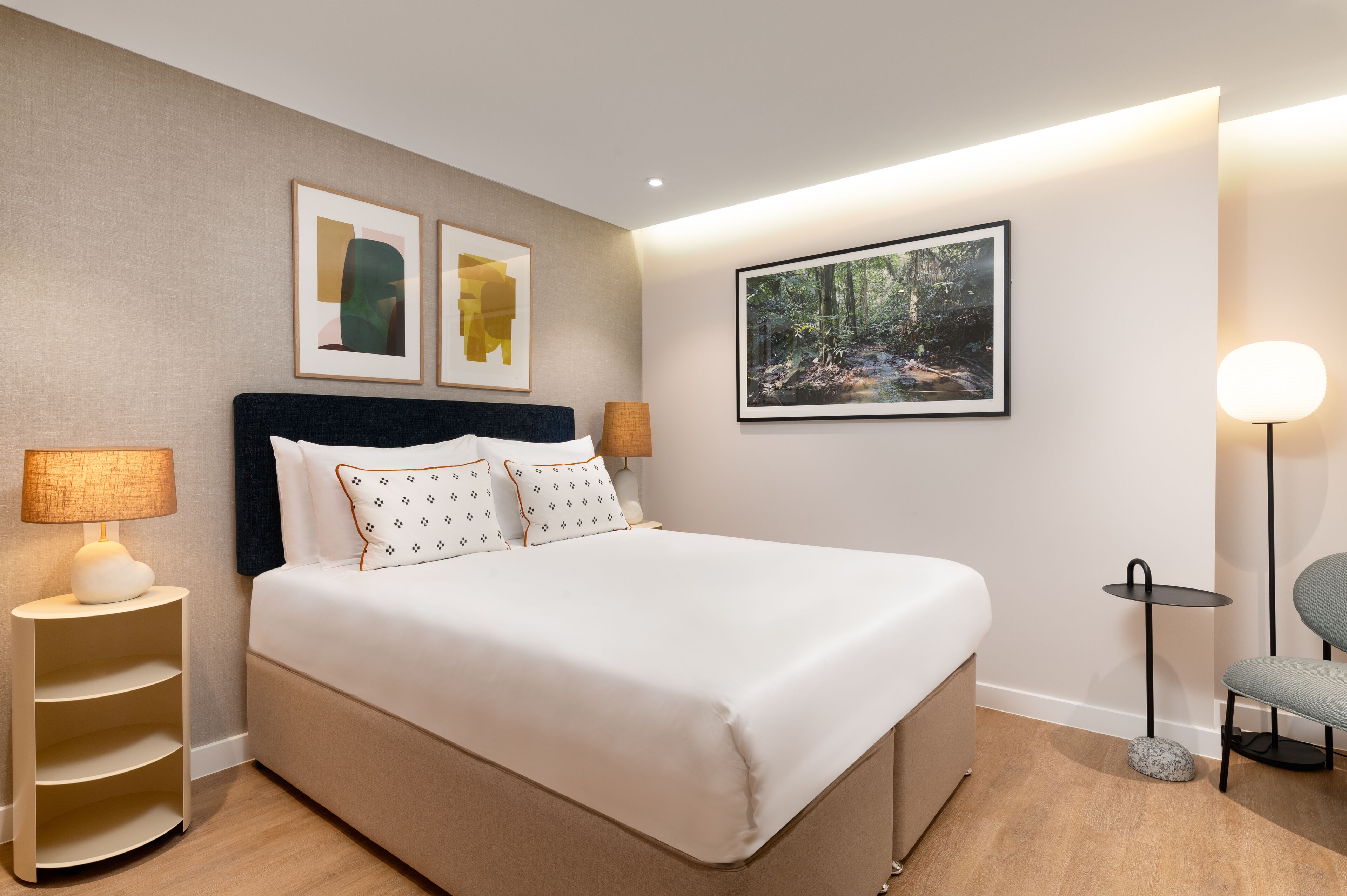 Un lit bien fait, avec une tête de lit, une table de chevet avec une lampe, un tableau encadré au mur et une petite table ronde avec une chaise.