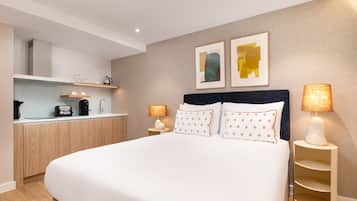 A modern hotel room with a large bed, bedside lamps, a small kitchenette, and two framed artworks on the wall.