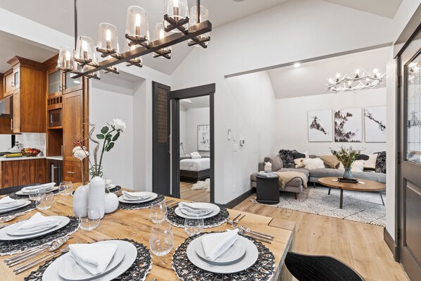 A modern dining area with a wooden table set for six, a chandelier, and a view into a bedroom.