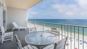 A balcony with a round glass table and white chairs, overlooking a beach with umbrellas and a clear view of the ocean.