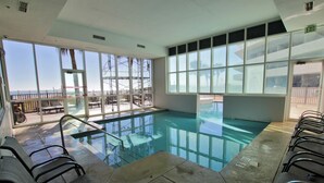 An indoor swimming pool with clear glass walls, a view of the beach, and several lounge chairs.