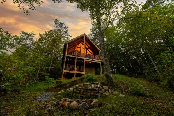 Uma cabana com varanda, cercada por árvores e um caminho de pedra que leva até ela.