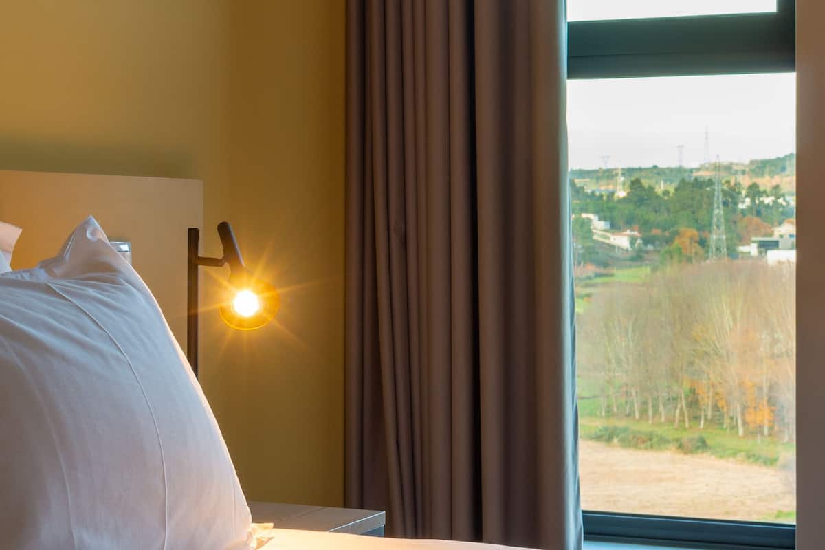 A hotel room with a bed, a nightstand, a lamp, and a window with curtains.