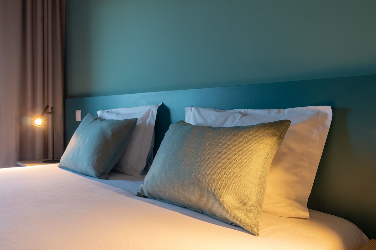 A neatly made bed with white linens and two pillows, a wall clock, and a curtain.