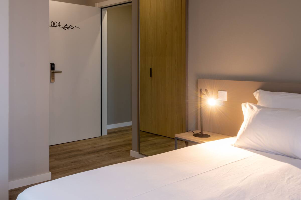 A hotel room with a bed, a nightstand with a lamp, a wooden panel wall, and a door with a number sign.