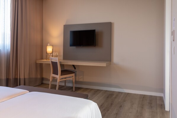 A hotel room with a bed, a desk with a chair, a flat-screen TV mounted on the wall, and a lamp on the desk. - Meu Hotel Porto Gandra (Paredes)
