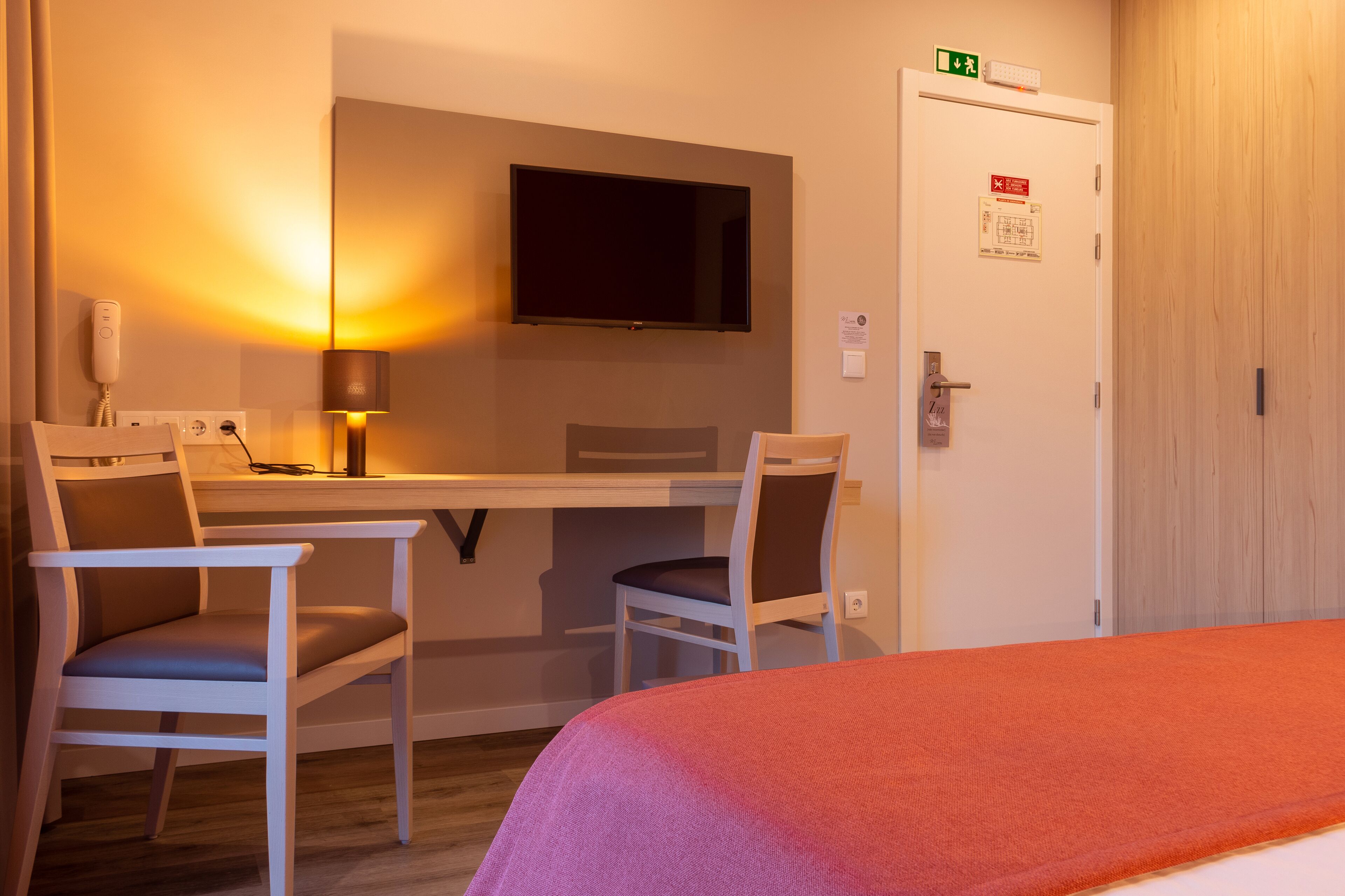 A hotel room with a bed, a desk with two chairs, a TV mounted on the wall, and an emergency exit sign.