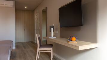 A modern room with a wooden floor, a desk with a chair, a flat-screen TV mounted on the wall, and a small table with fruits and a book.