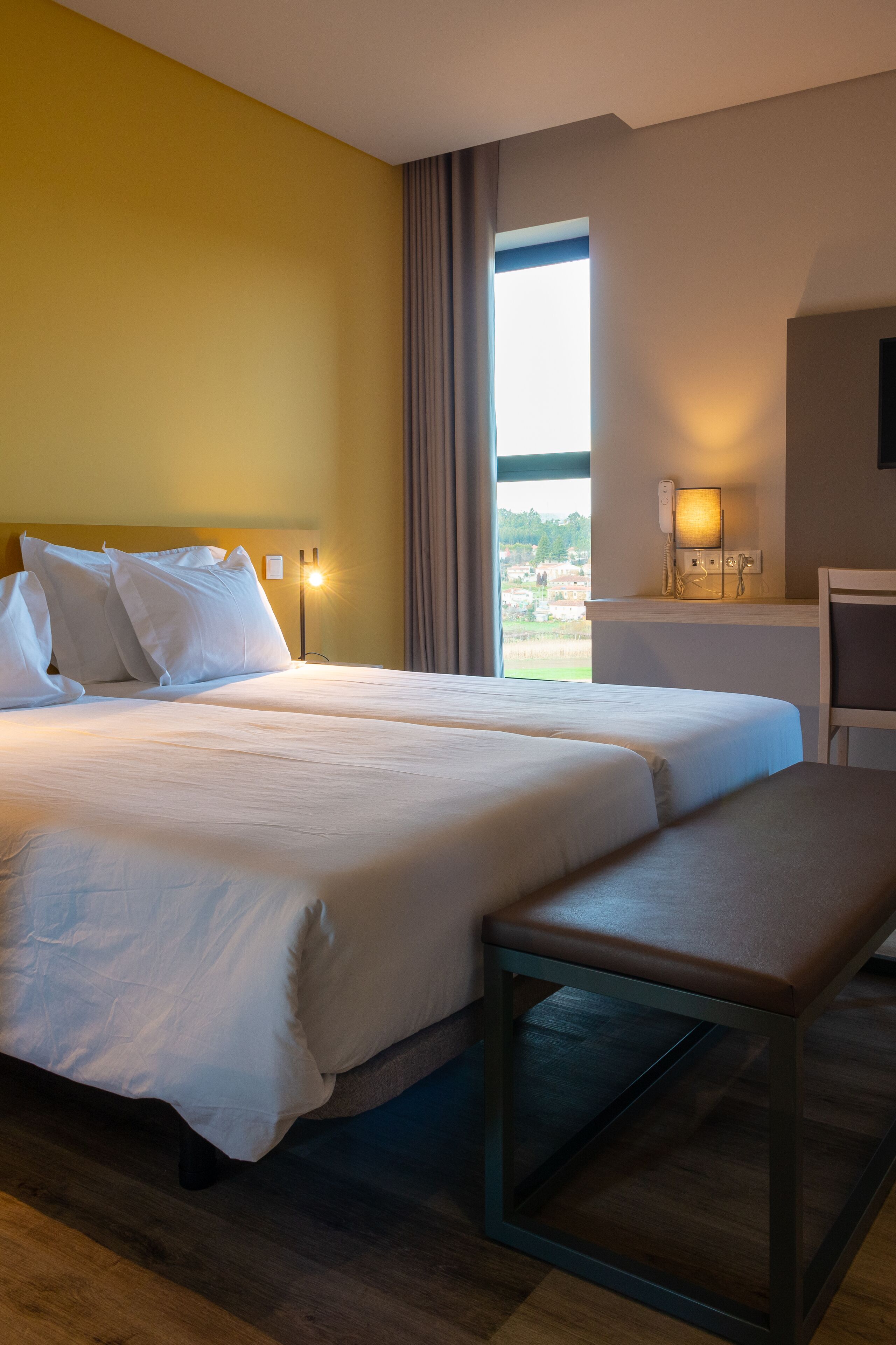 A hotel room with two beds, a desk, and a large window offering a view.