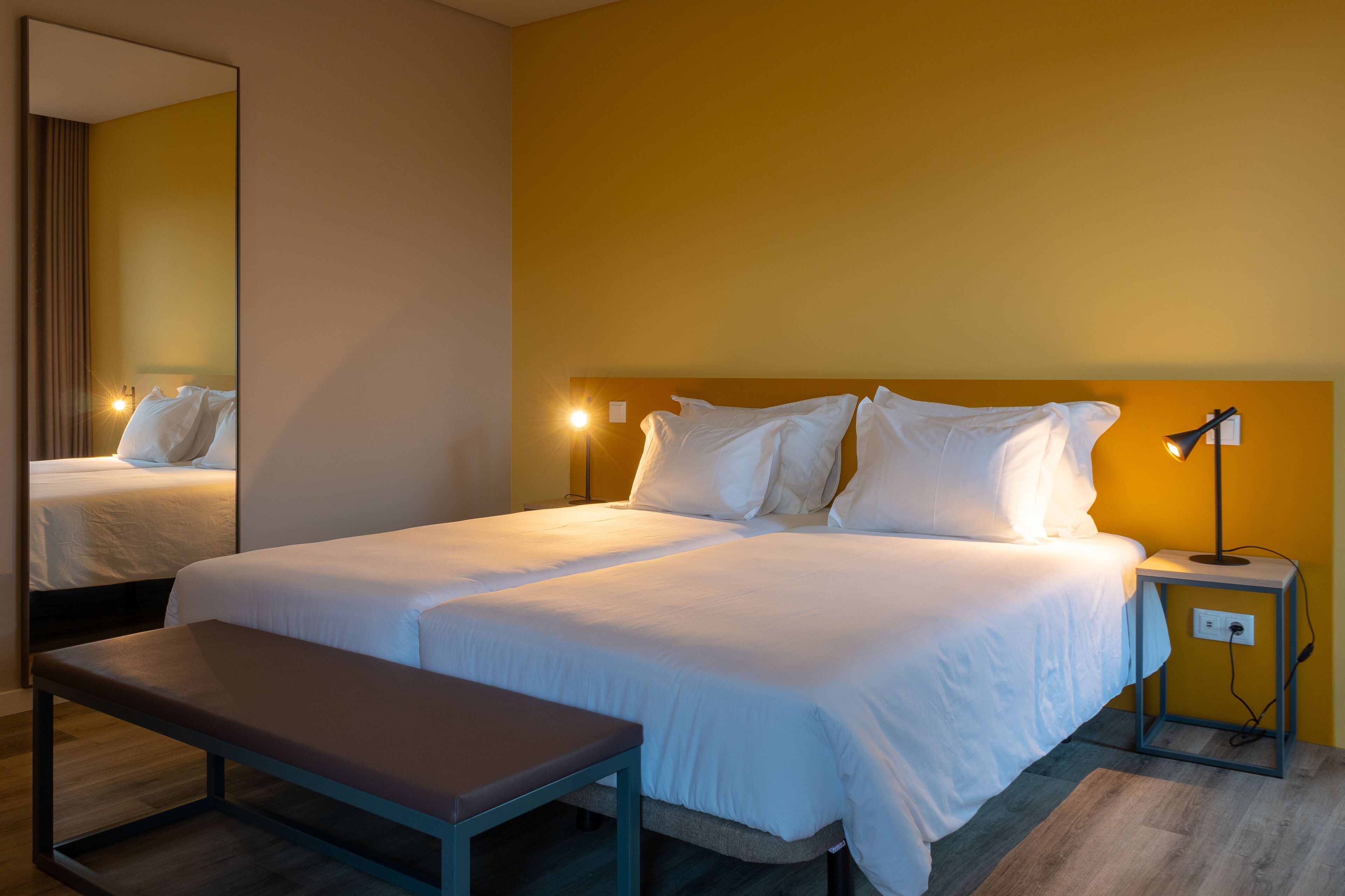 A hotel room with a large bed, a bedside table, a bench, and a nightstand with a lamp.