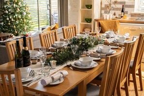 A dining table set for a meal with chairs, a Christmas tree, and a kitchen in the background.