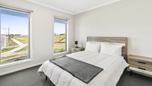 A bedroom with a large bed, two bedside tables, and two windows with views of a grassy area and a road. - Relax at Torquay Dunes: Shops and Beaches Nearby (Torquay)