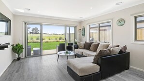 A modern living room with a sofa, a coffee table, and a view through glass doors to a green landscape. - Relax at Torquay Dunes: Shops and Beaches Nearby (Torquay)