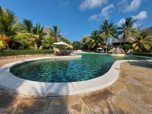 A clear pool with lounge chairs and umbrellas, surrounded by palm trees and thatched-roof structures. - Tembo Court Apartment (Malindi)