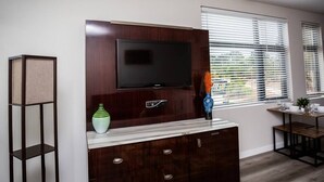 A modern living room with a flat-screen TV mounted on a dark wood cabinet, a small side table with a vase, and a window with blinds.