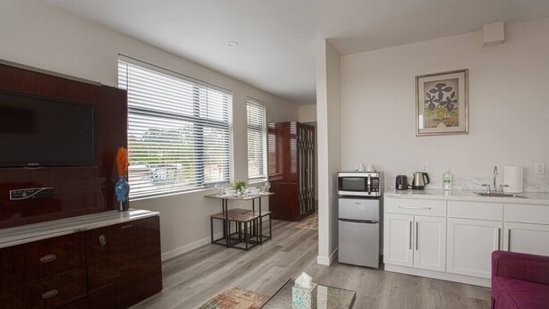 A modern living room with a flat-screen TV, a wooden cabinet, a coffee table, and a kitchen area with a refrigerator, microwave, and sink.