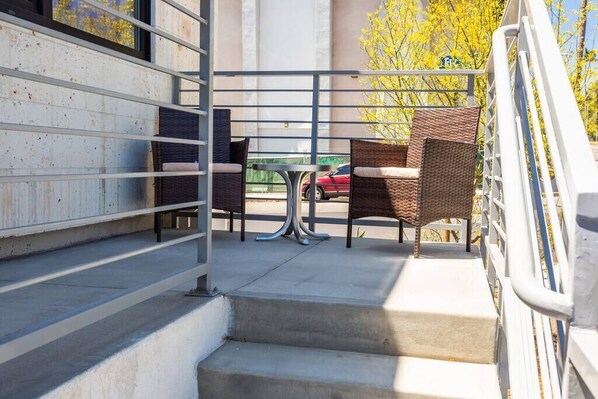 A balcony with wicker chairs and a table, a staircase, and a view of a red car and yellow trees.