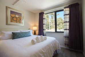 A neatly made bed with white linens and rolled towels, a framed artwork on the wall, a bedside lamp, and a window with purple curtains.