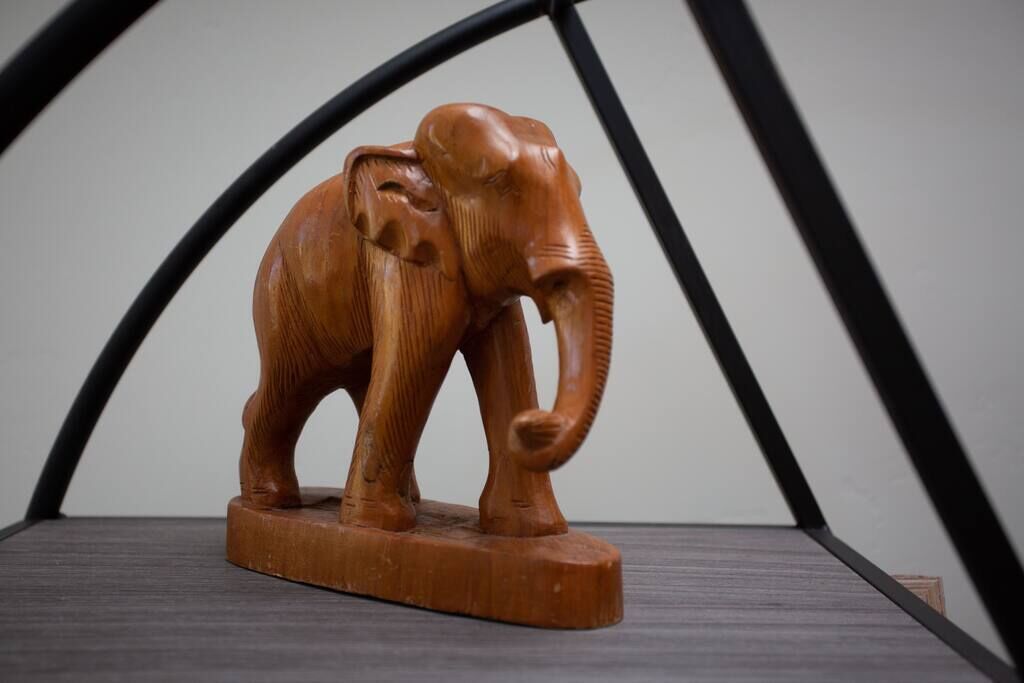 A wooden elephant sculpture on a dark wooden base, placed on a chair with a black metal frame.
