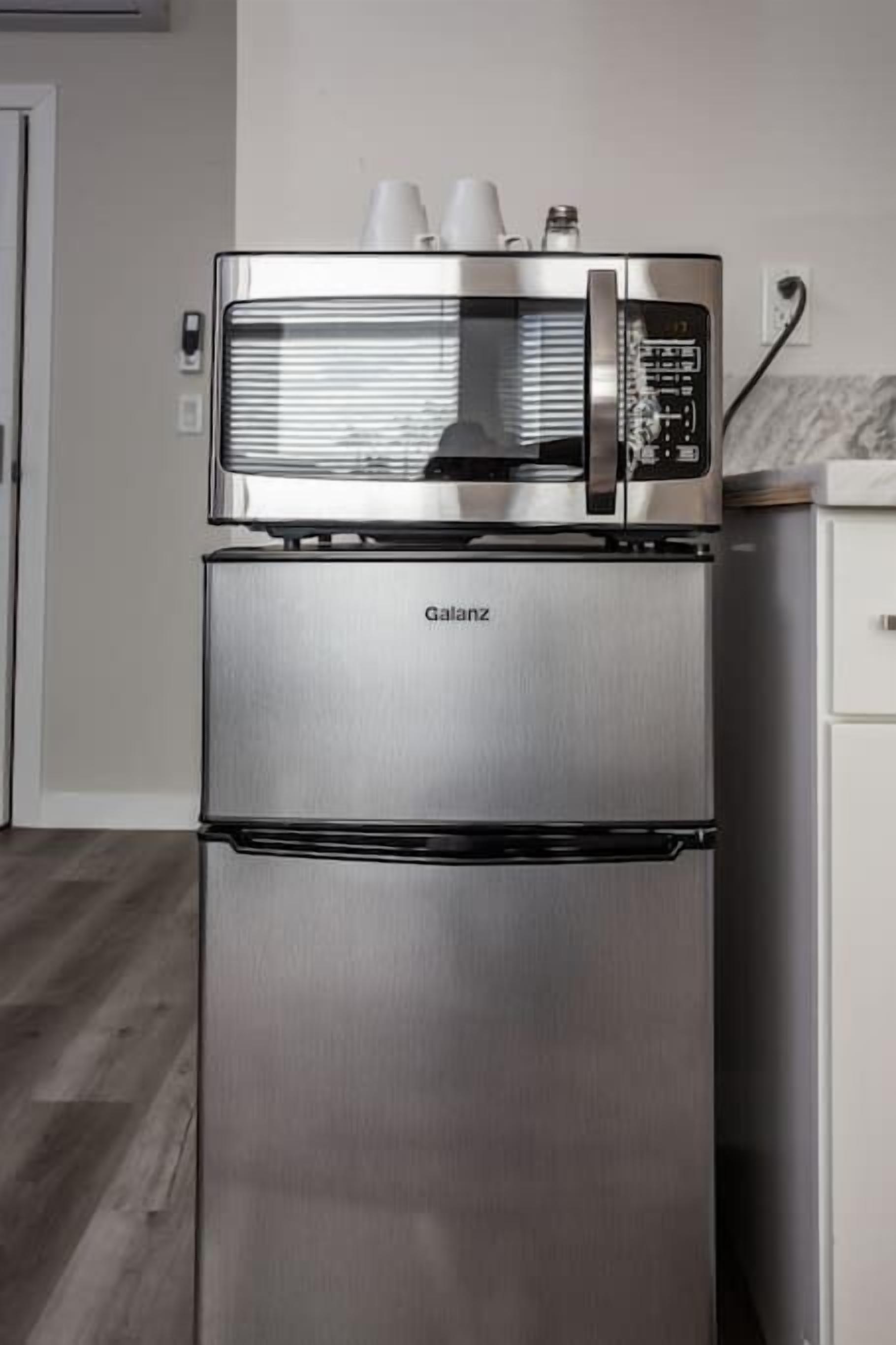 A stainless steel Galanz refrigerator with a built-in microwave on top, located in a kitchen with a wooden floor and white cabinetry.