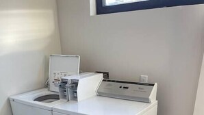 Een kleine wasruimte met een voorlader wasmachine en droger, een klein aanrecht met een koffiezetapparaat, en een raam boven de droger.