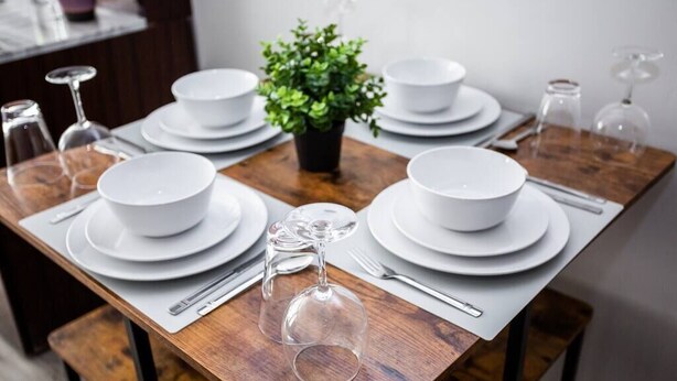 Een eettafel gedekt met witte borden, kommen, bestek en wijnglazen, met een bloempot in het midden.