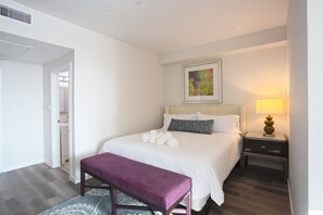 A hotel room with a bed, a bench, a nightstand, a lamp, and a framed picture on the wall.