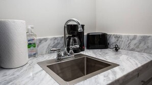Et badeværelse med vask i rustfrit stål, toiletpapir, håndspritdispenser og en sort kaffemaskine.