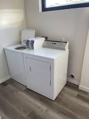 A compact laundry room with a front-loading washer and dryer, a small countertop with a water dispenser, and a window above the dryer.