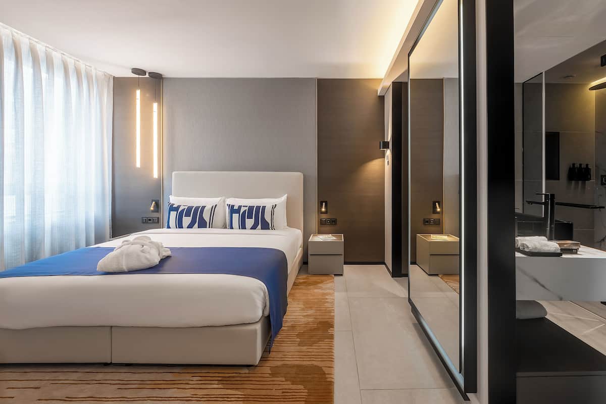 A modern hotel room with a large bed, bedside tables, a mirror, and a bathroom visible through a glass partition.