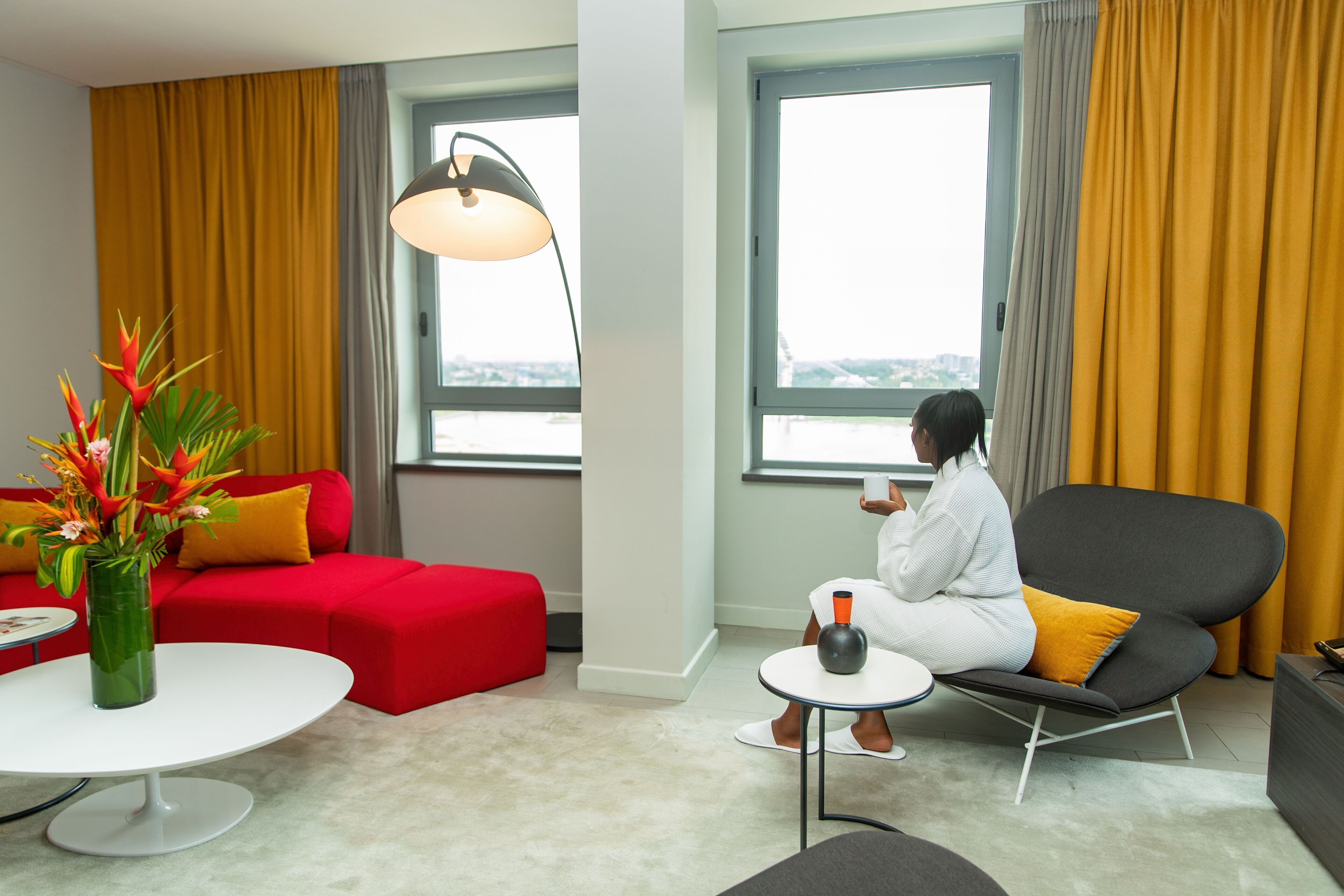 Une chambre d’hôtel moderne avec un canapé rouge, un fauteuil noir, une table basse blanche, une lampe sur pied et de grandes fenêtres avec des rideaux.