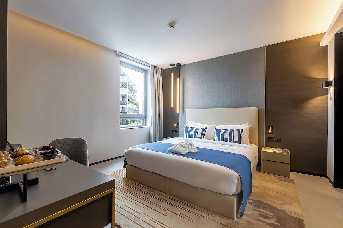 A modern hotel room with a large bed, a desk with breakfast items, a window with a view of buildings, and a wall-mounted light fixture.