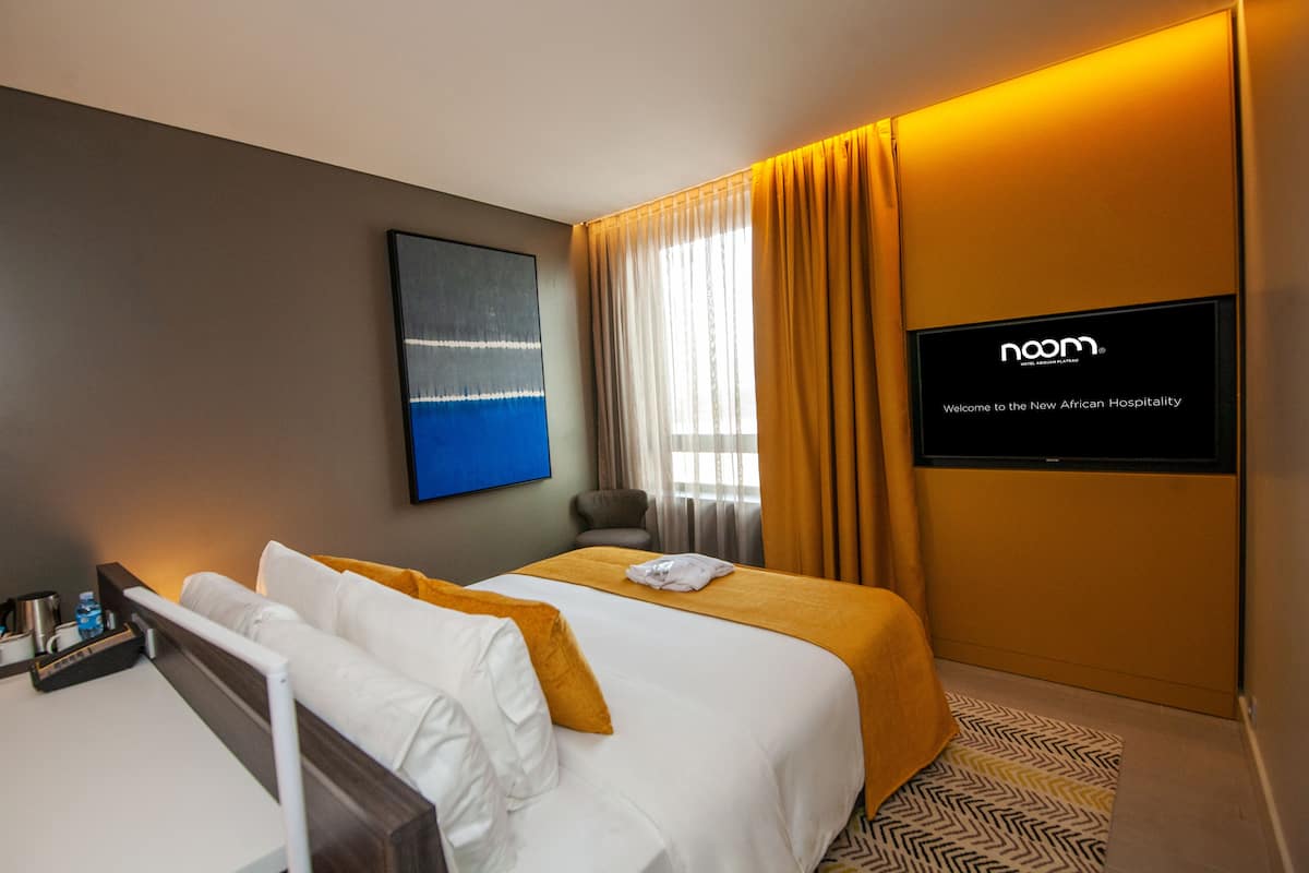 A hotel room with two beds, a TV screen displaying 'noam Welcome to the New African Hospitality', a painting on the wall, and a window with curtains.