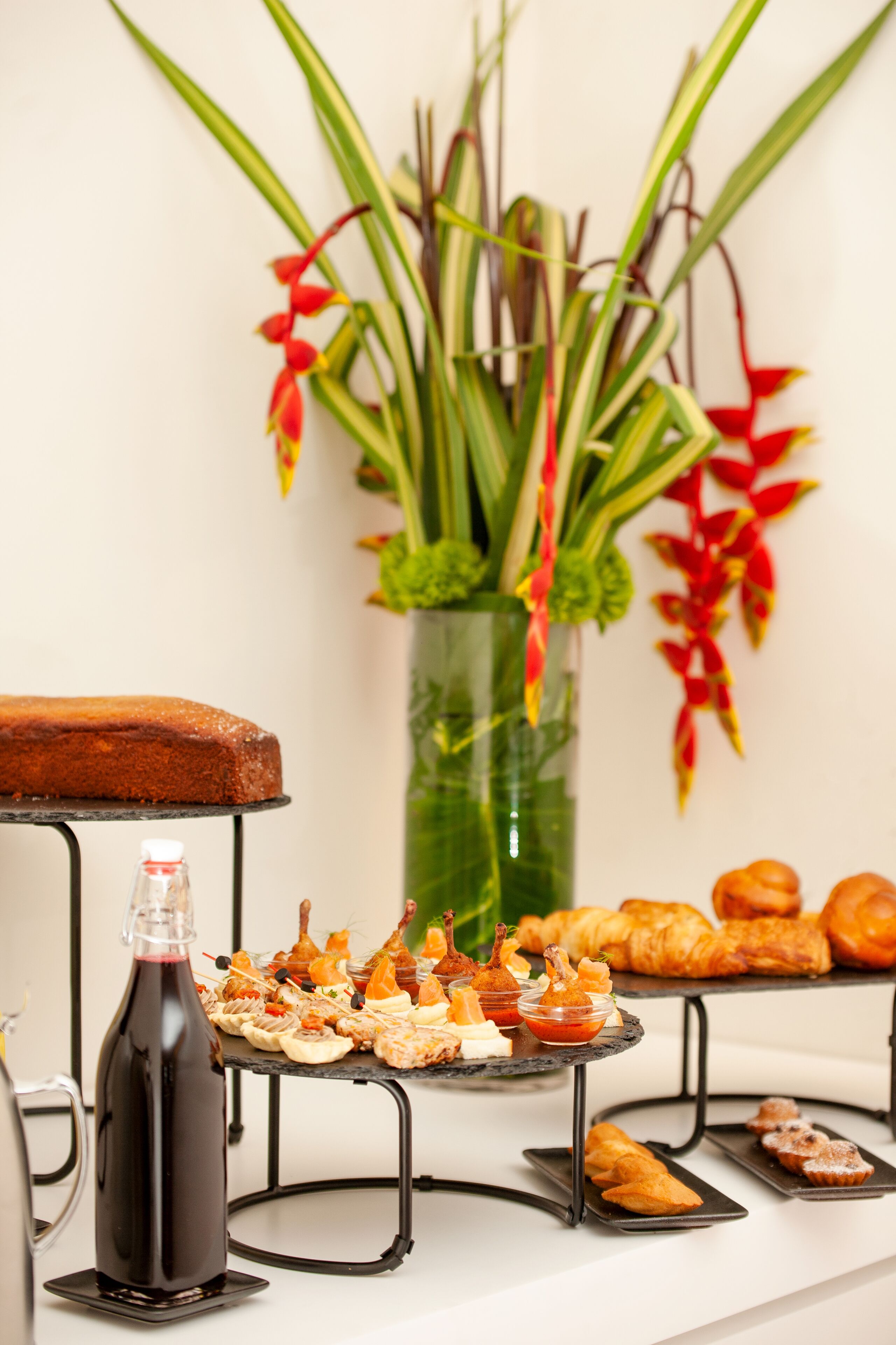 A buffet table with various food items, a bottle of red beverage, and a vase with green and red flowers.