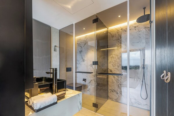 Un baño moderno con ducha envuelta en vidrio, pared de azulejos de mármol y lavamanos empotrado con toallero.
