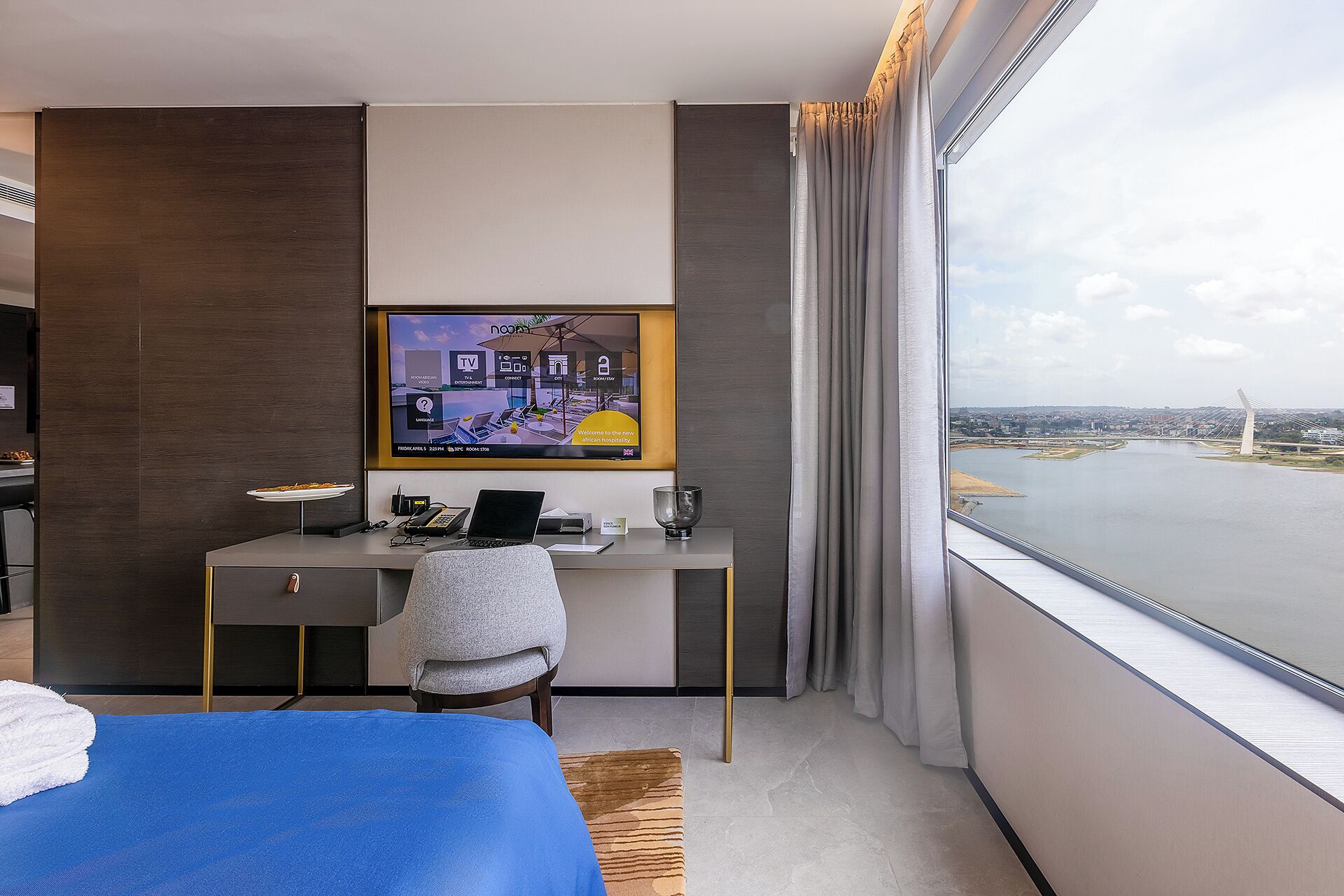 Una habitación de hotel con un ventanal grande, un escritorio con una computadora portátil, un televisor y una cama.
