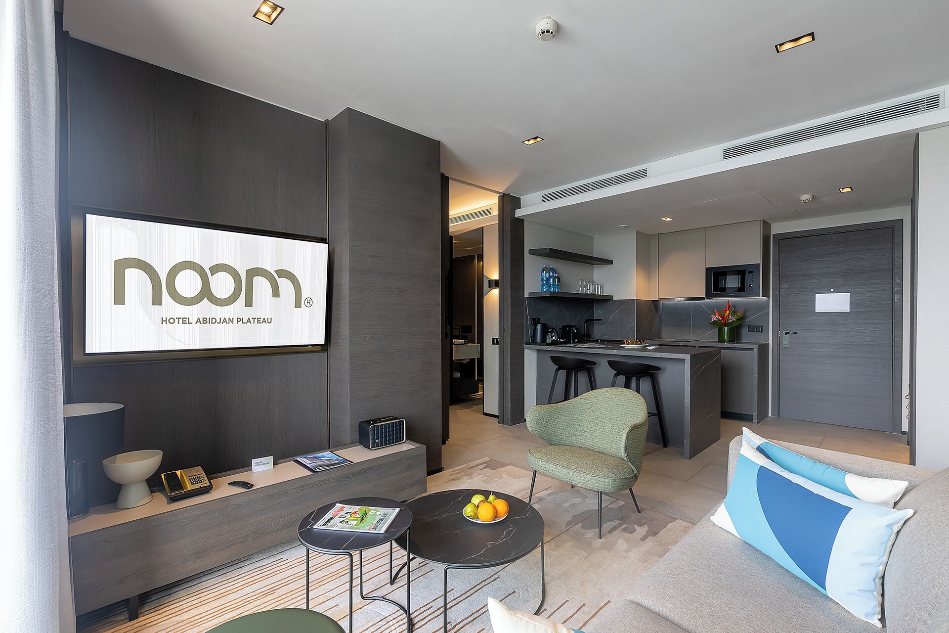 A modern hotel room with a dark wood finish, a flat-screen TV, a small table with a bowl and remote, a green armchair, and a sofa with blue cushions.