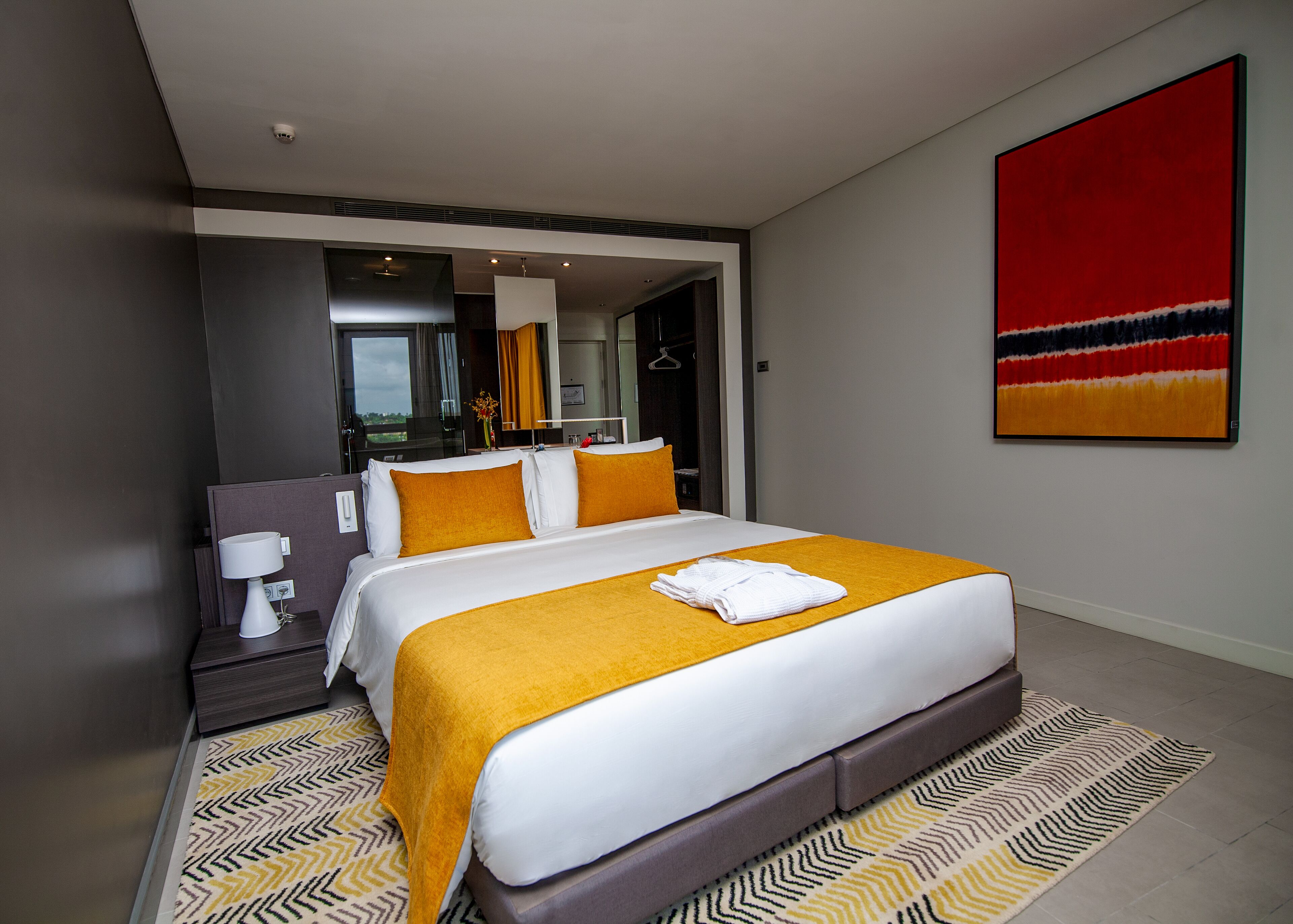 A modern hotel room with a large bed, a bedside table with a lamp, a mirror, and a red abstract painting on the wall.