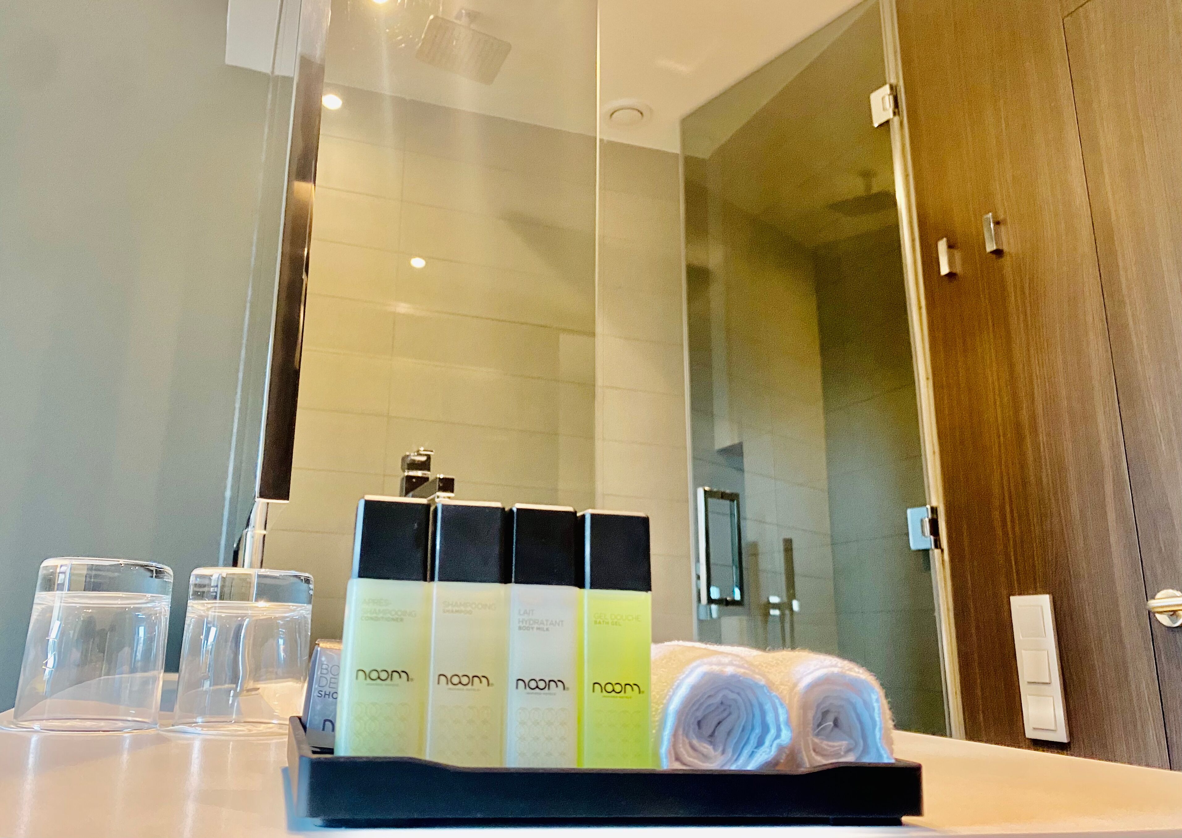 A bathroom with a glass shower enclosure, a wooden door, and a counter with toiletries including shampoo and body wash.