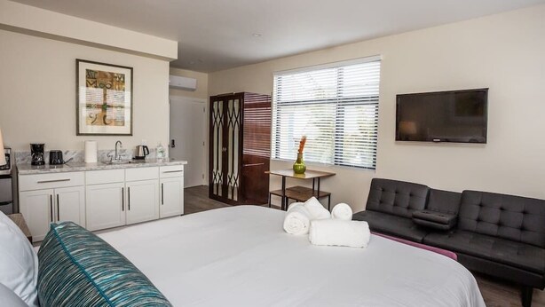 Ein Hotelzimmer mit Bett, einer Couch, einem Fernseher, einem Schreibtisch mit Stuhl, einem Fenster mit Jalousien und einer Küchenzeile.