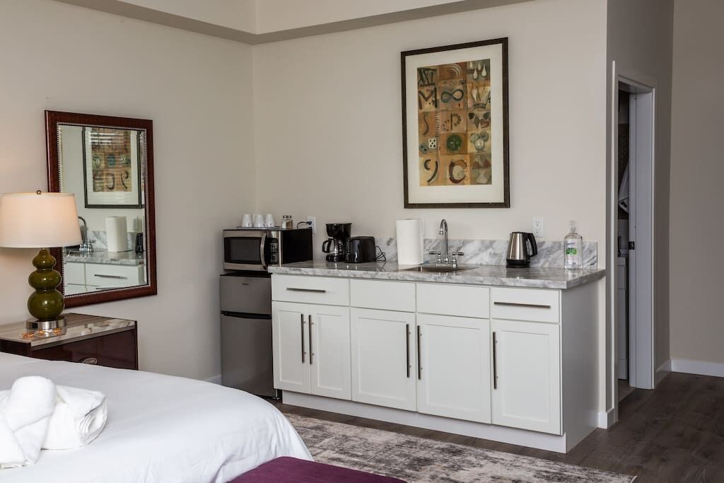 Une chambre d’hôtel comprenant un lit, un bureau avec une lampe, une kitchenette équipée d’une machine à café et d’un four à micro-ondes, ainsi qu’une œuvre d’art encadrée accrochée au mur.