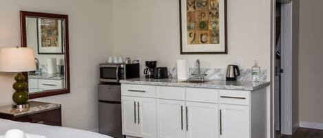 Habitación de hotel con cama, escritorio con lámpara, kitchenette con cafetera y microondas, además de un cuadro en la pared.