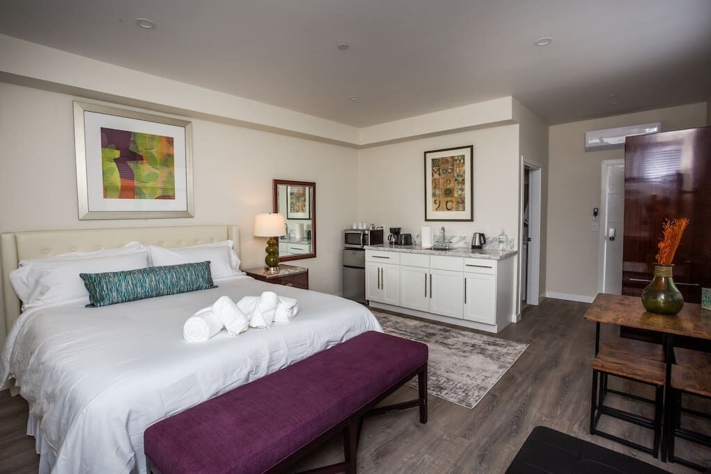Quarto de hotel com cama, um banco, mesa de jantar, área de cozinha e obras de arte nas paredes.