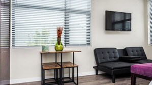 Ein modernes Wohnzimmer mit einer schwarzen Couch, einem kleinen Holztisch mit einer Vase und einem an der Wand befestigten Fernseher.