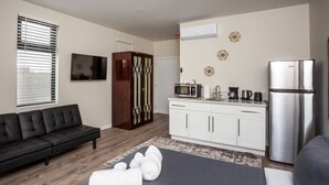 Een moderne woonkamer met een zwarte bank, een eethoek met witte kasten, een keuken met een koelkast en een flatscreen televisie aan de muur.