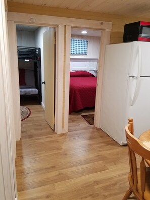 Une petite pièce comprenant un lit superposé, un lit simple avec une couverture rouge, un réfrigérateur blanc et une table en bois.