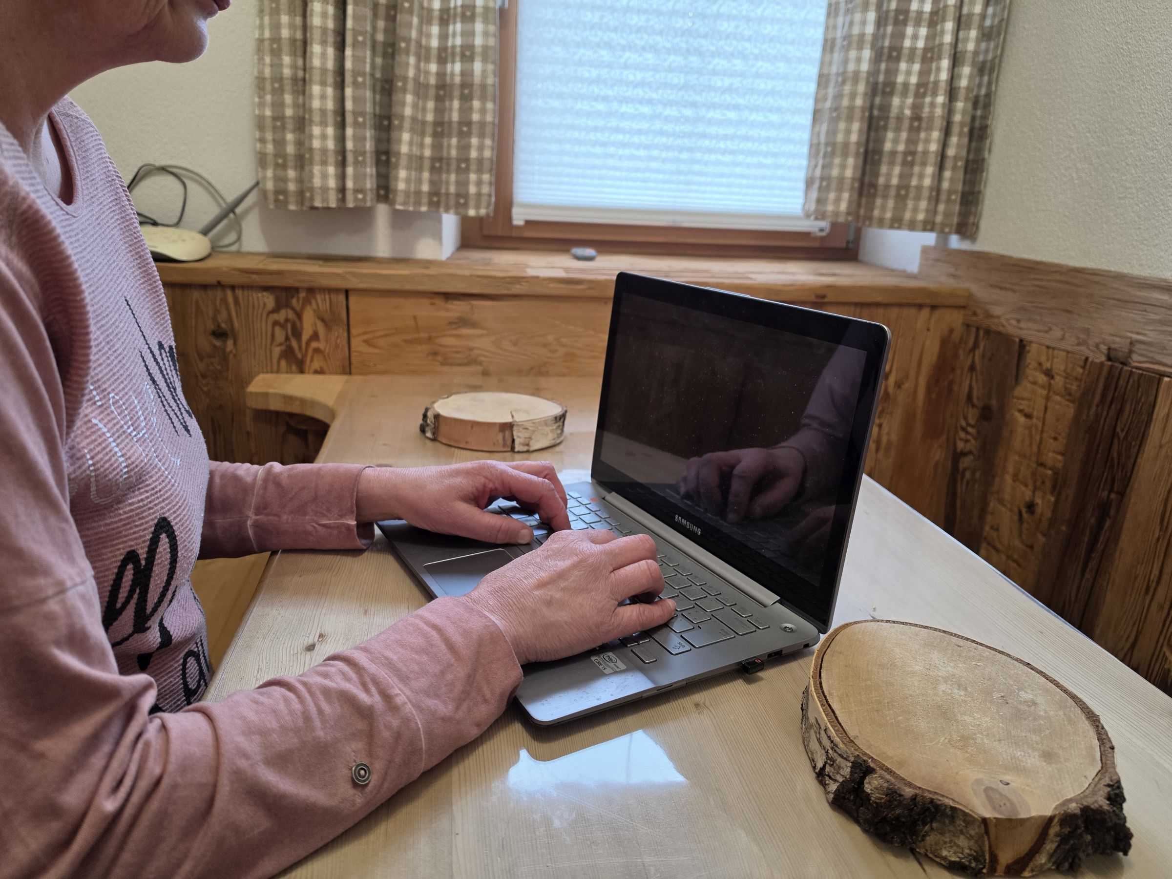 A person using a laptop on a wooden table with a wooden bench and a window with curtains.