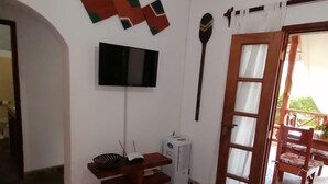A room with a wooden table, a TV mounted on the wall, a ceiling fan, and a door leading to an outdoor area with a chair. - Johari Villas (Malindi)