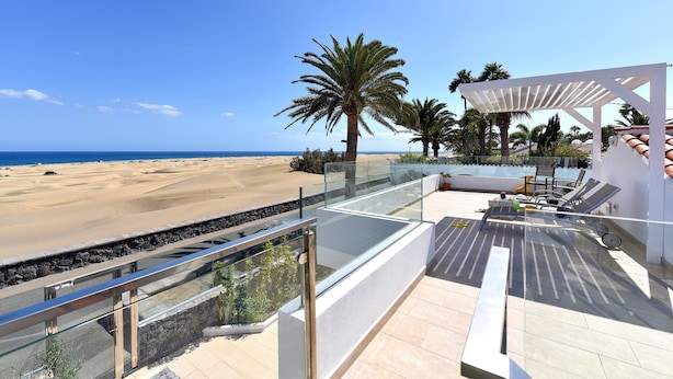 Una terraza en la azotea con barandilla de cristal que da a una playa, palmeras y una pérgola blanca con asientos.